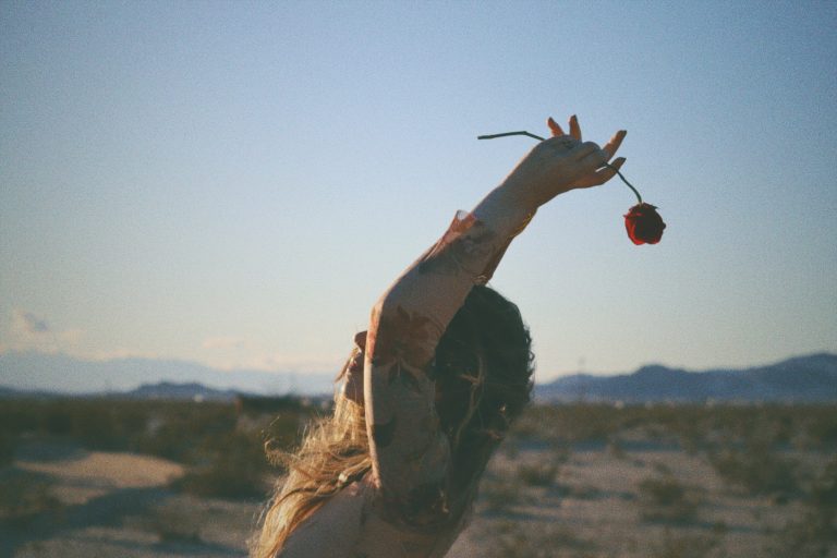 Empowered woman raising arms to the sky with a rose backdrop, symbolizing liberation, confidence, and becoming her next-level self.